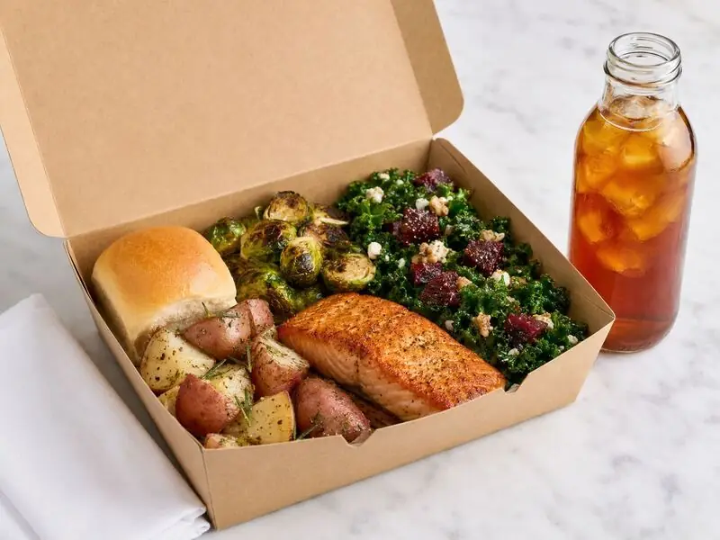 SF Catering Company office meal box containing roasted salmon, colorful greens, root vegetables, bread, and a refreshing iced drink.